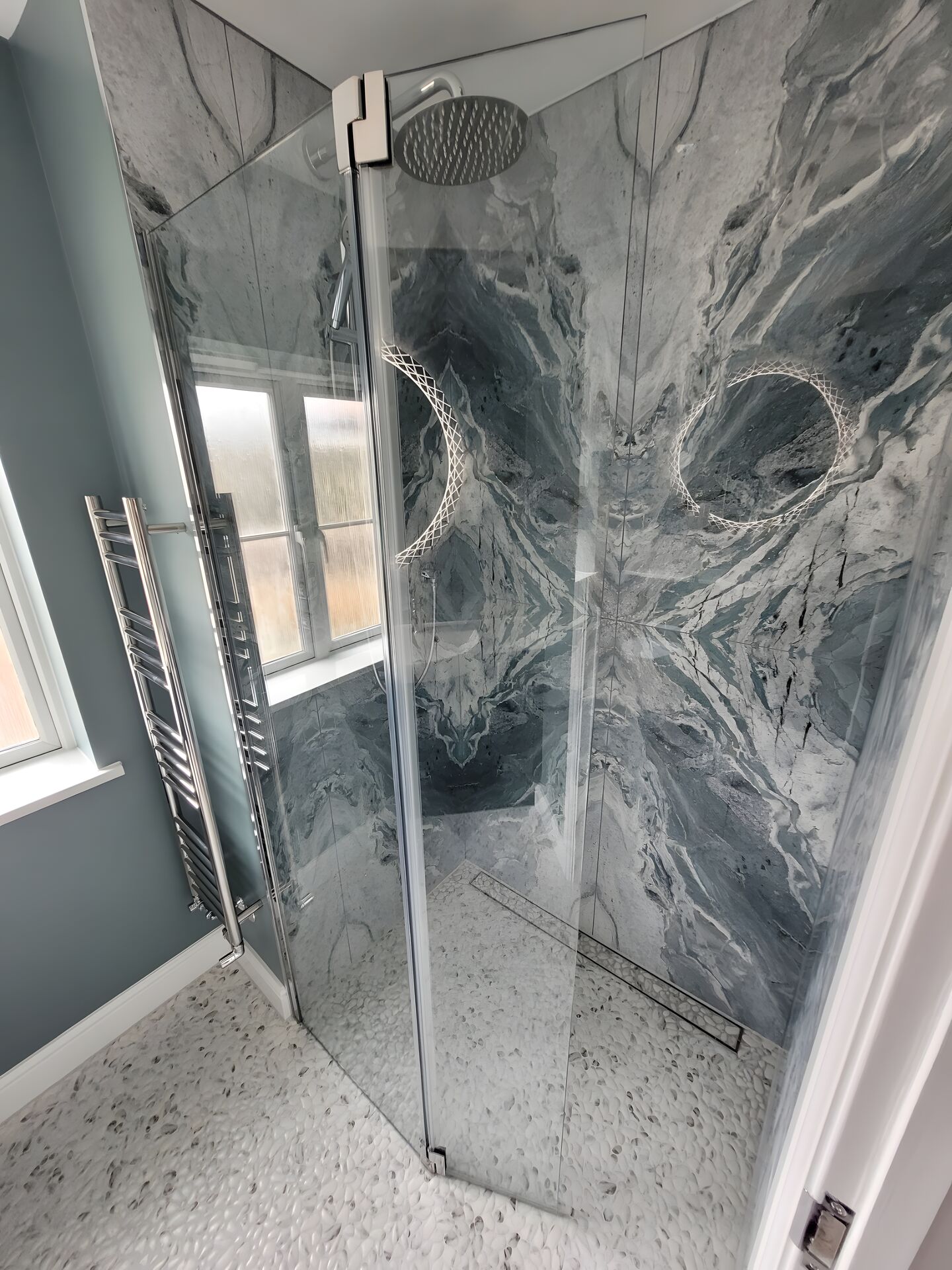 Walk-in shower lined with blue-grey marble-effect porcelain slabs, the veining mirrored across the joins into a starburst, over a white pebble-effect floor with a linear drain