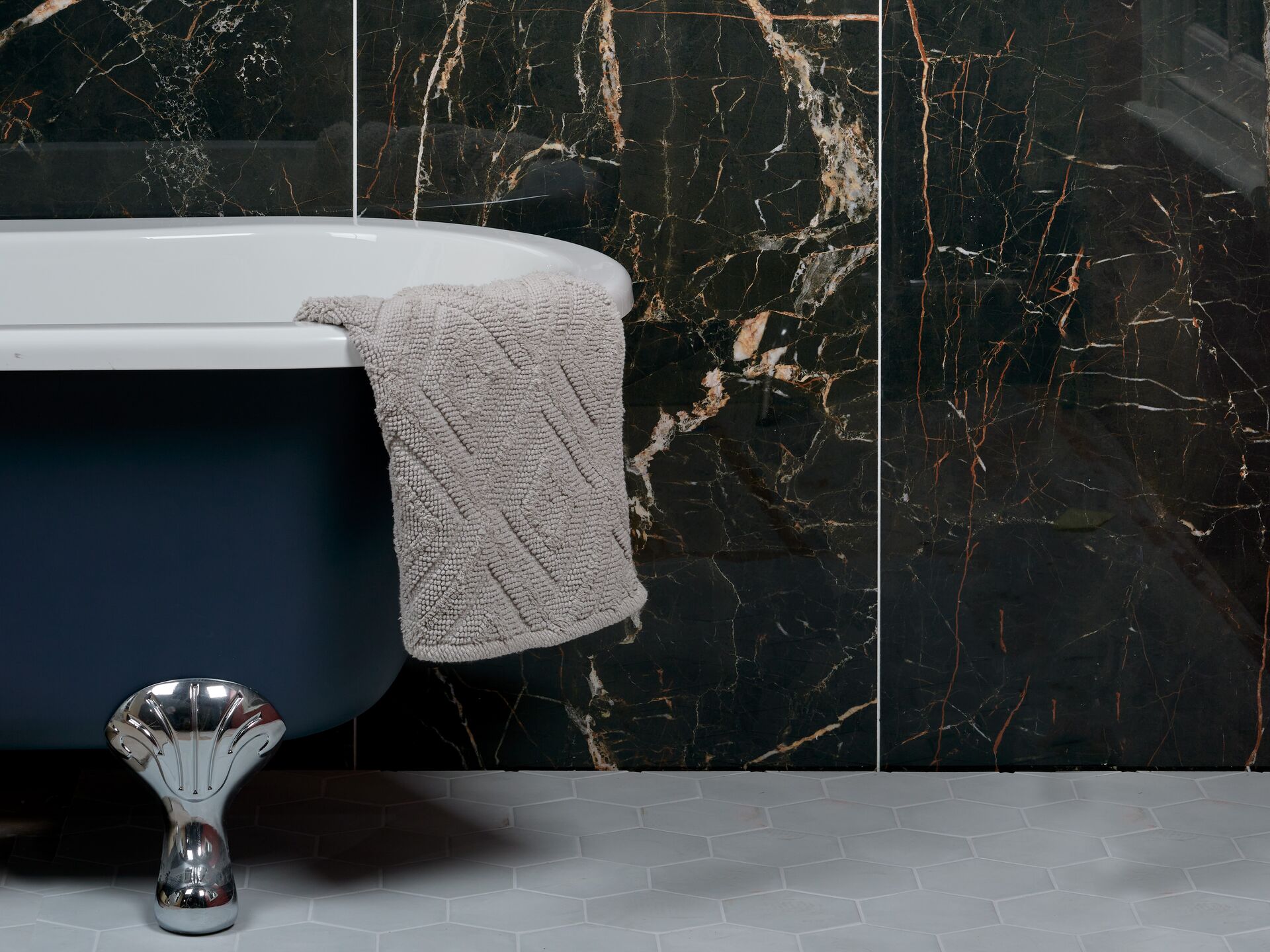 Black marble-effect porcelain wall tiles with copper and white veining, divided by two thin pale grout joints, behind a navy roll-top bath on a grey hexagon tile floor