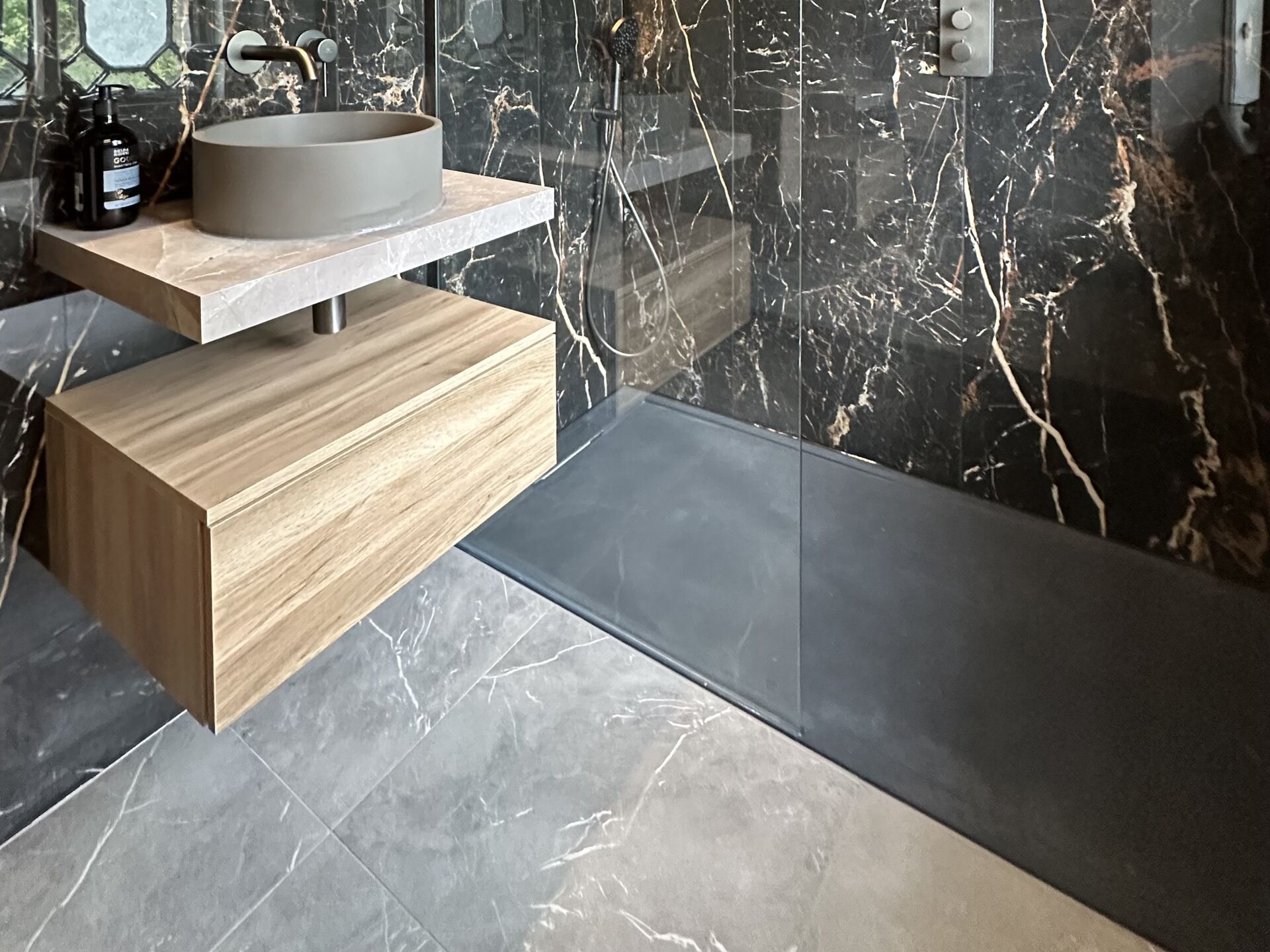 Large grey marble-effect porcelain floor tiles with daylight reflecting across them, beside a dark shower tray, an oak wall-hung drawer unit and black marble-effect wall tiles