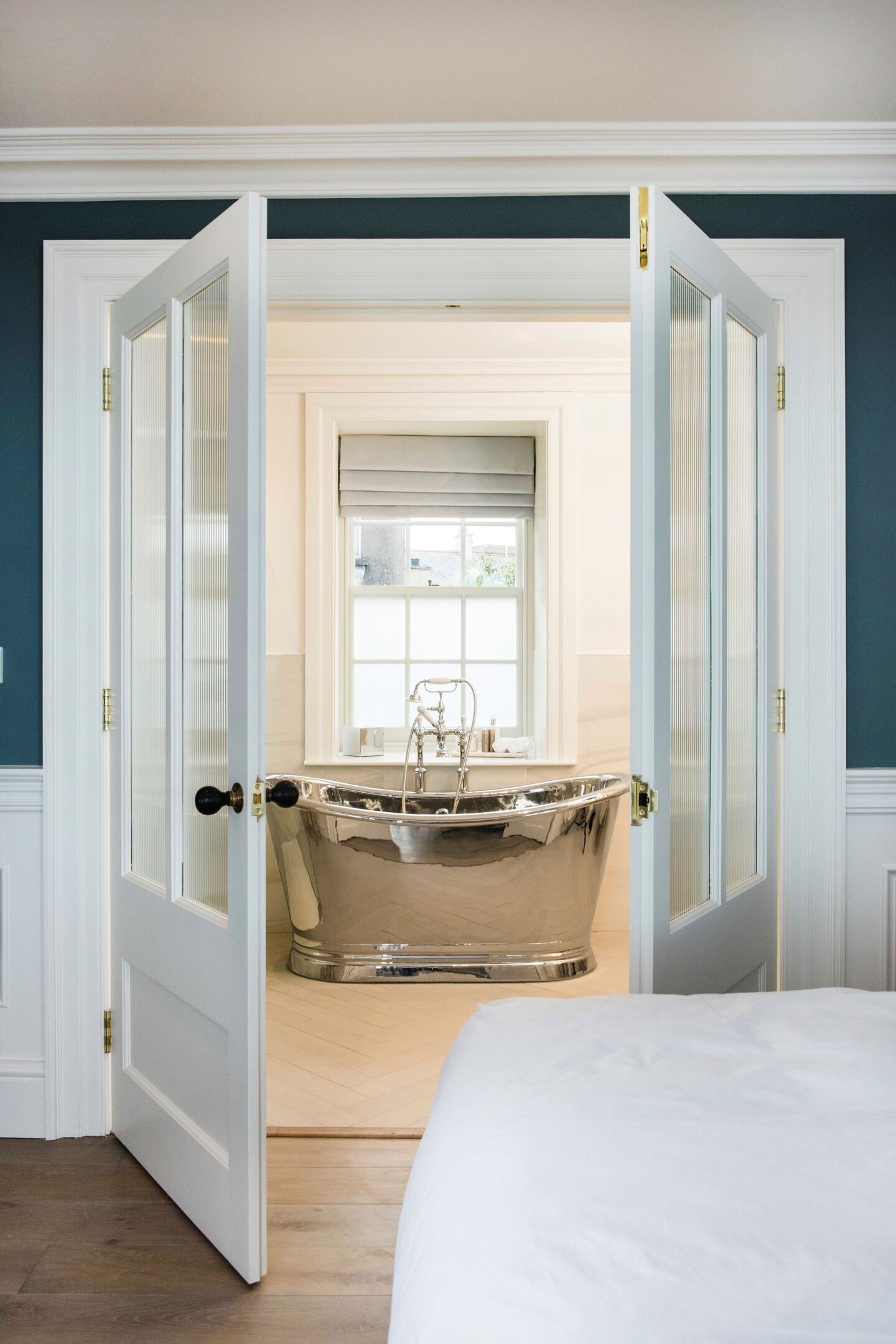 Nickel-finish freestanding bath seen through open glazed double doors from a bedroom, with the bed corner in the foreground