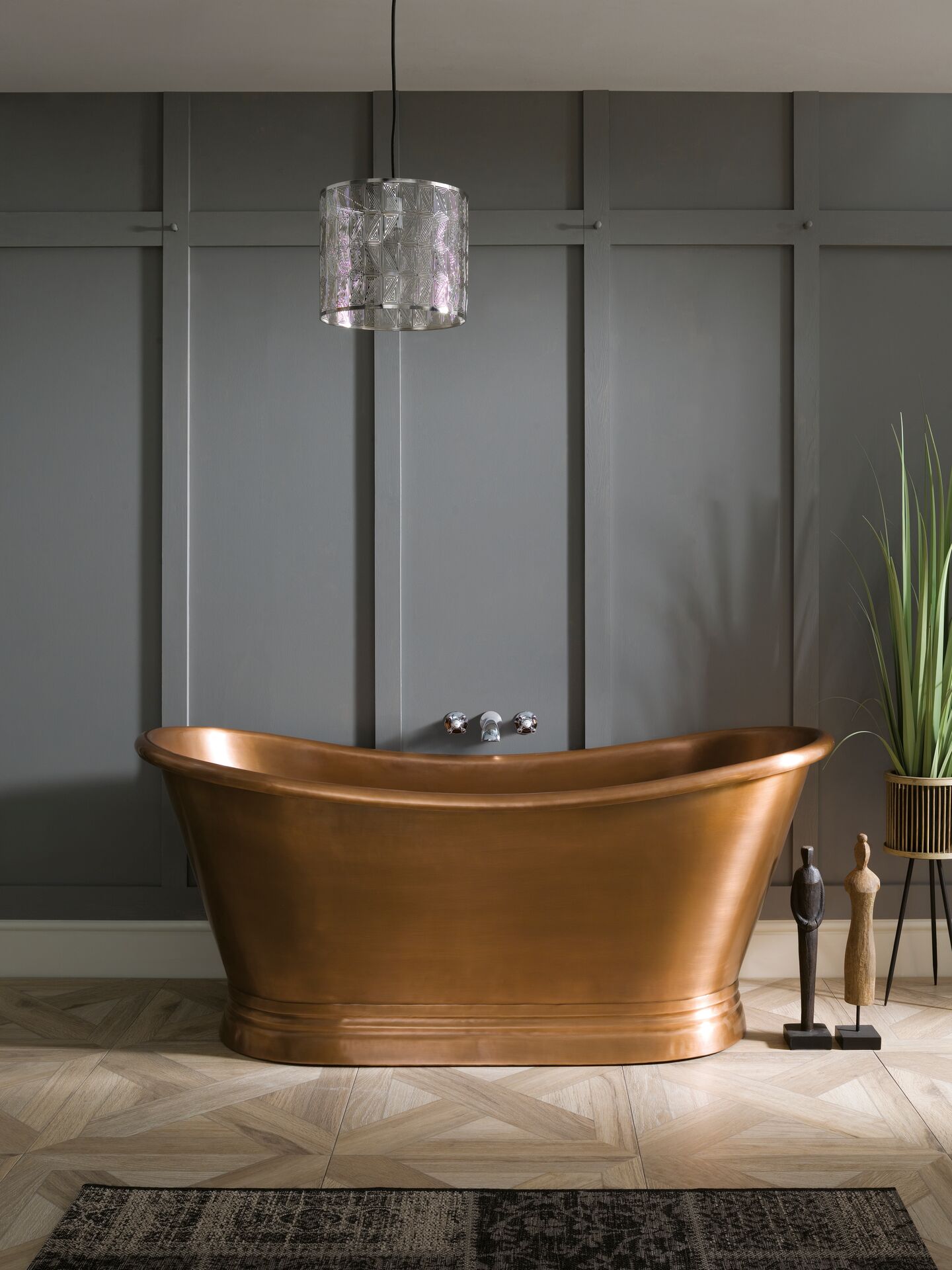 BC Designs Copper Boat Bath standing on a wood-effect parquet floor laid in a diamond pattern, against a panelled grey wall, with a rug in front