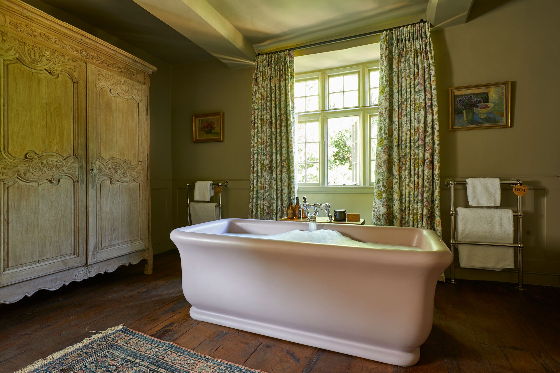 BC Designs freestanding bath filled with bubbles on a dark wood floor, beside a carved oak armoire and in front of a curtained window, with a chrome heated towel rail either side