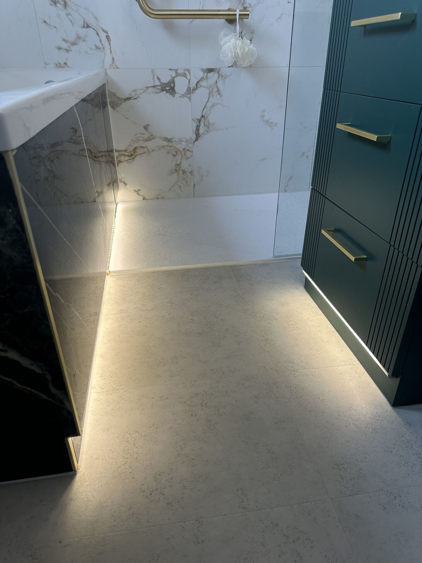 A warm strip in a slim channel along the base of a tiled bath panel where it meets the floor, and a second strip under an emerald drawer unit