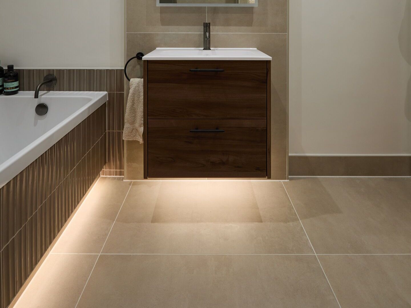 Floating walnut vanity with a warm line of light on the floor beneath it and a second warm glow along the base of a fluted-tile bath panel, with no strip visible