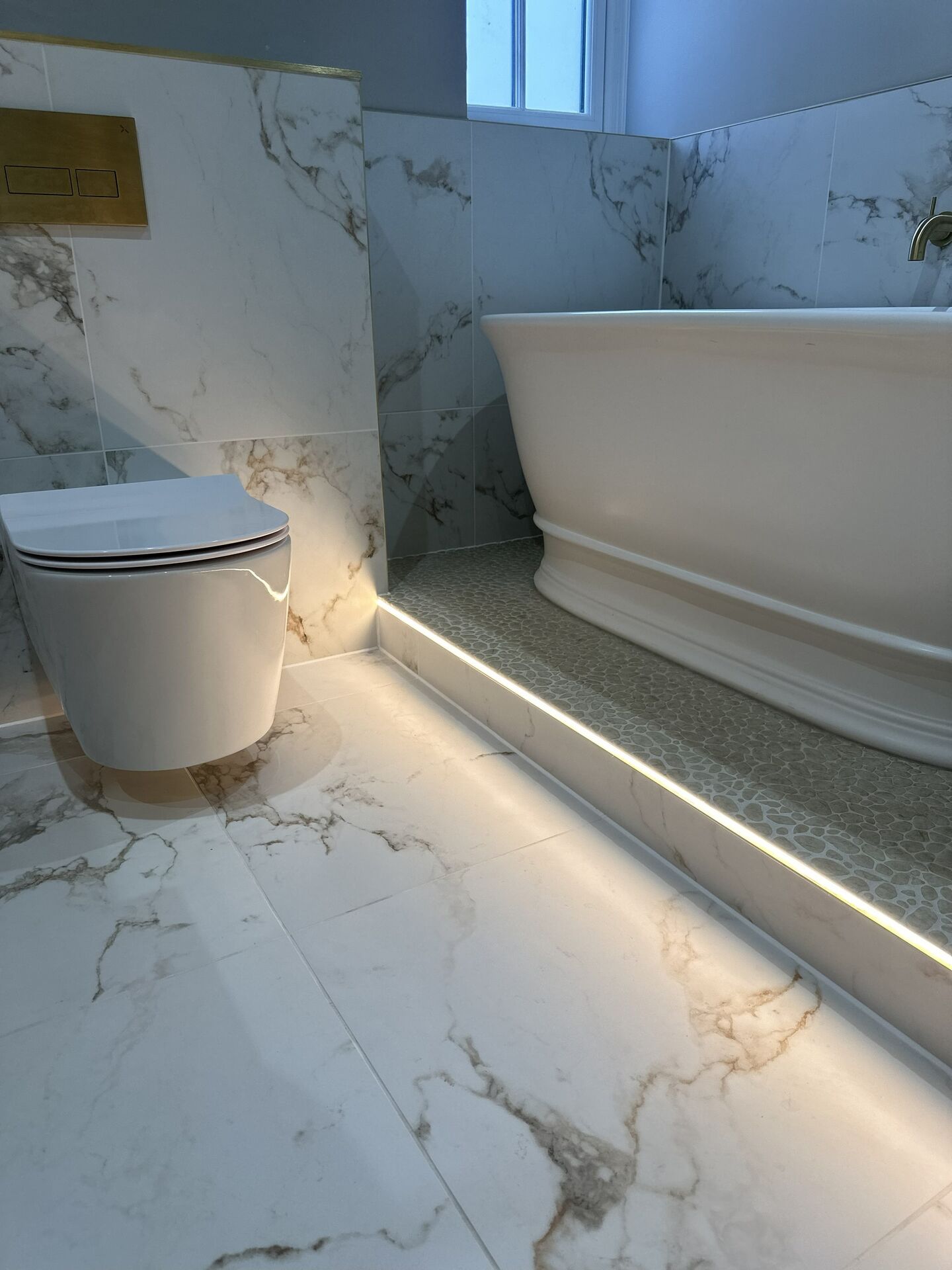 A warm continuous strip tucked under the nosing of a raised pebble-mosaic bath ledge, lighting the marble-effect floor beside a wall-hung WC