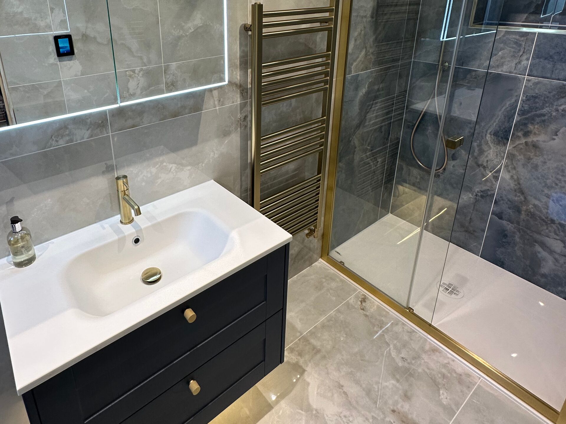Master ensuite in Wickham Bishops, Essex: a navy wall-hung vanity unit with a white basin and brushed brass tap, a brushed brass towel radiator, and a brushed brass framed shower enclosure with a white tray, all on grey marble-effect tiles