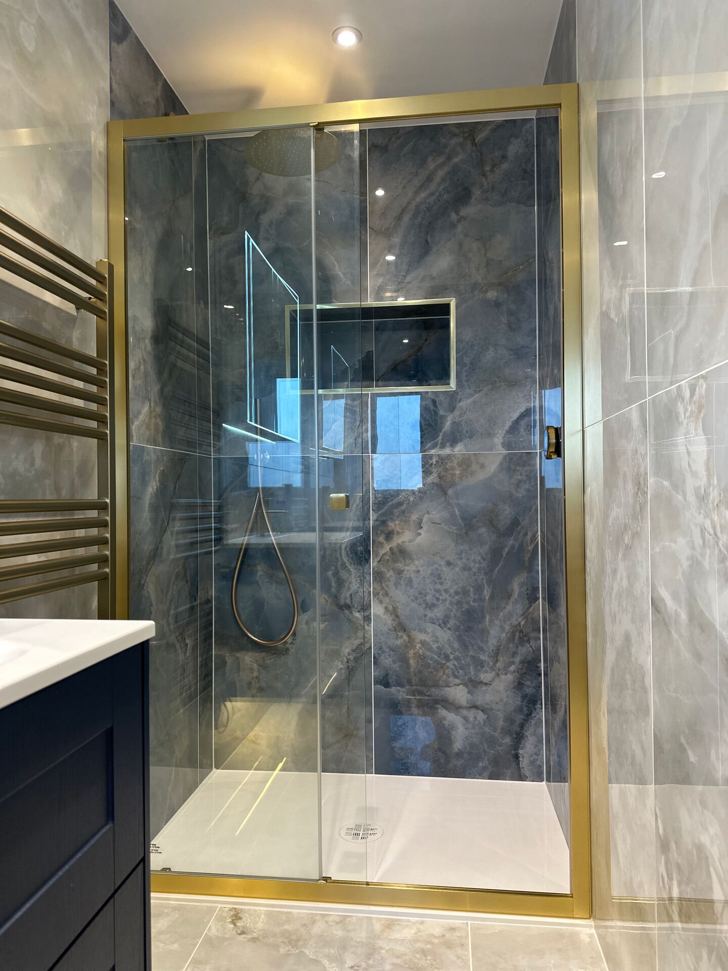 Brushed brass framed shower enclosure across the end of the Wickham Bishops ensuite, with a dark blue-grey marble-effect tiled back wall, a brass-framed niche and a rain shower head, the corner of the navy vanity unit in the foreground and a brushed brass towel radiator on the left