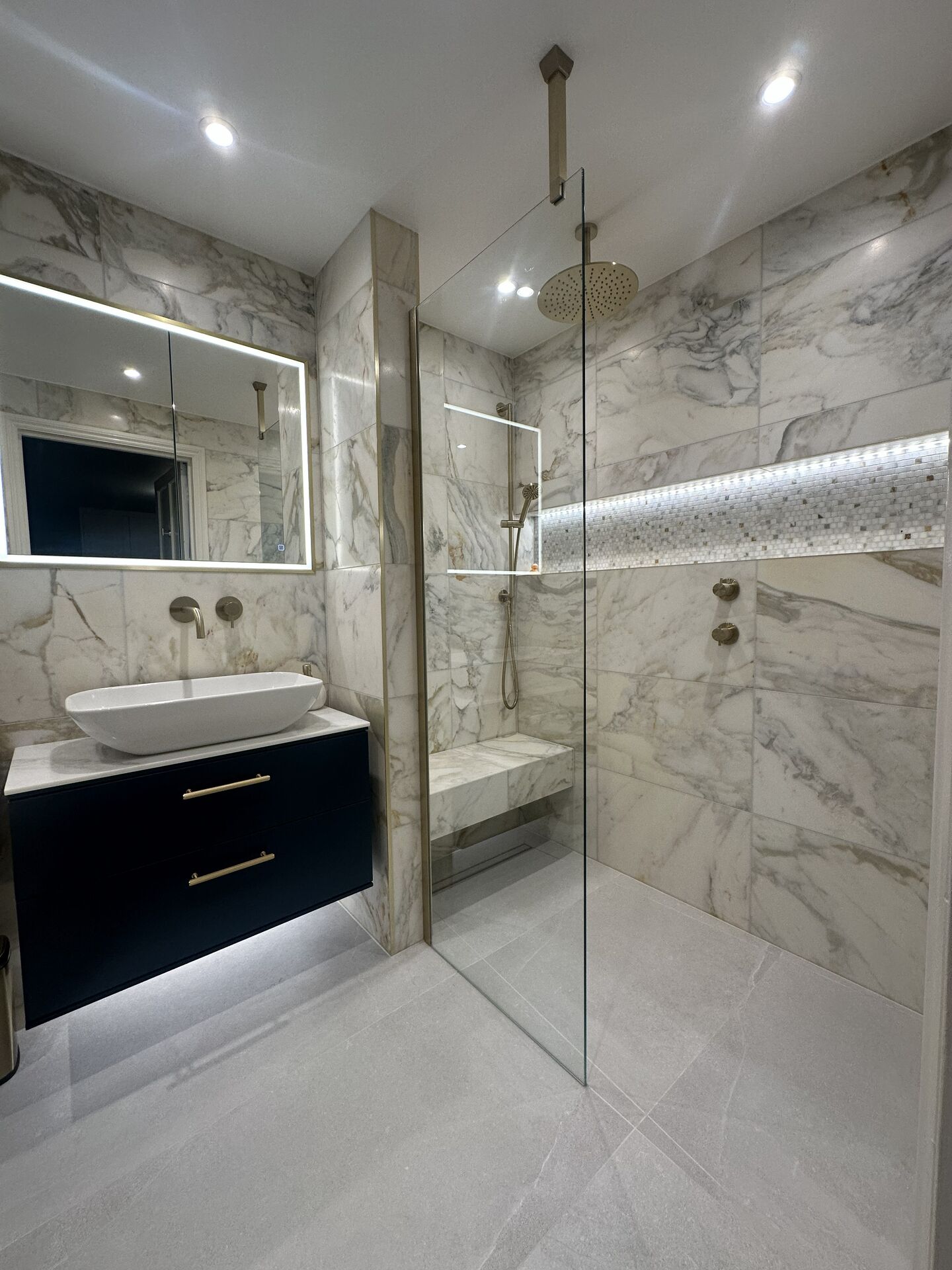 The finished Chelmsford CM3 master ensuite: a navy wall-hung vanity unit with a white vessel basin and wall-mounted tap under an LED mirrored cabinet, veined marble walls, a glass shower screen, a ceiling rain head, a tiled bench and a lit mosaic niche strip