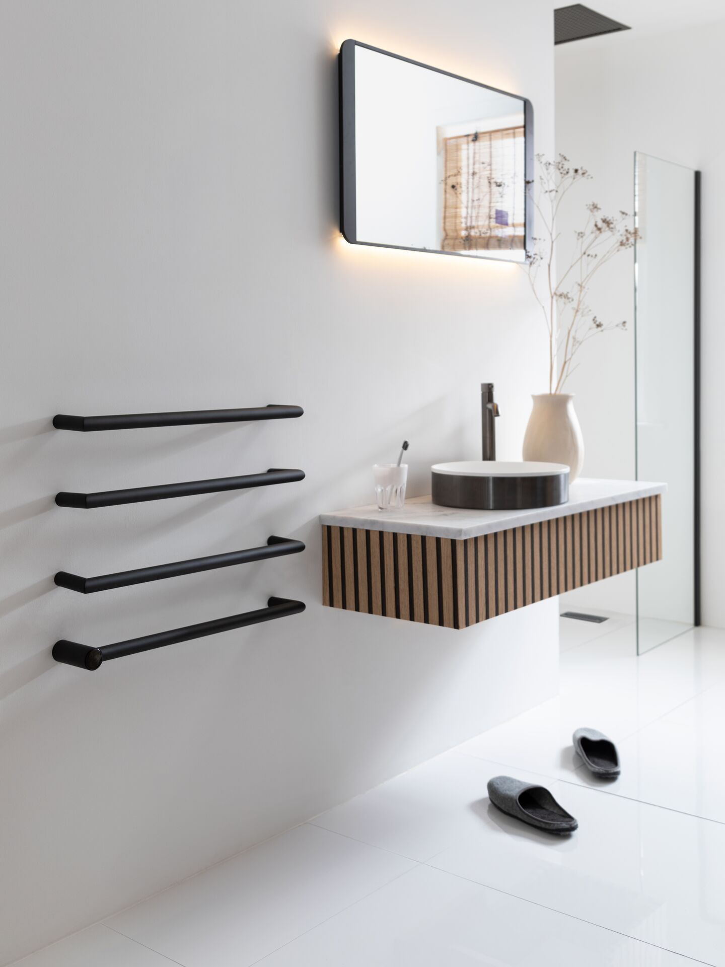 Black JTP Obi towel radiator on a white wall beside a wall-hung vanity with a vessel basin, a glass shower screen beyond and slippers on the floor