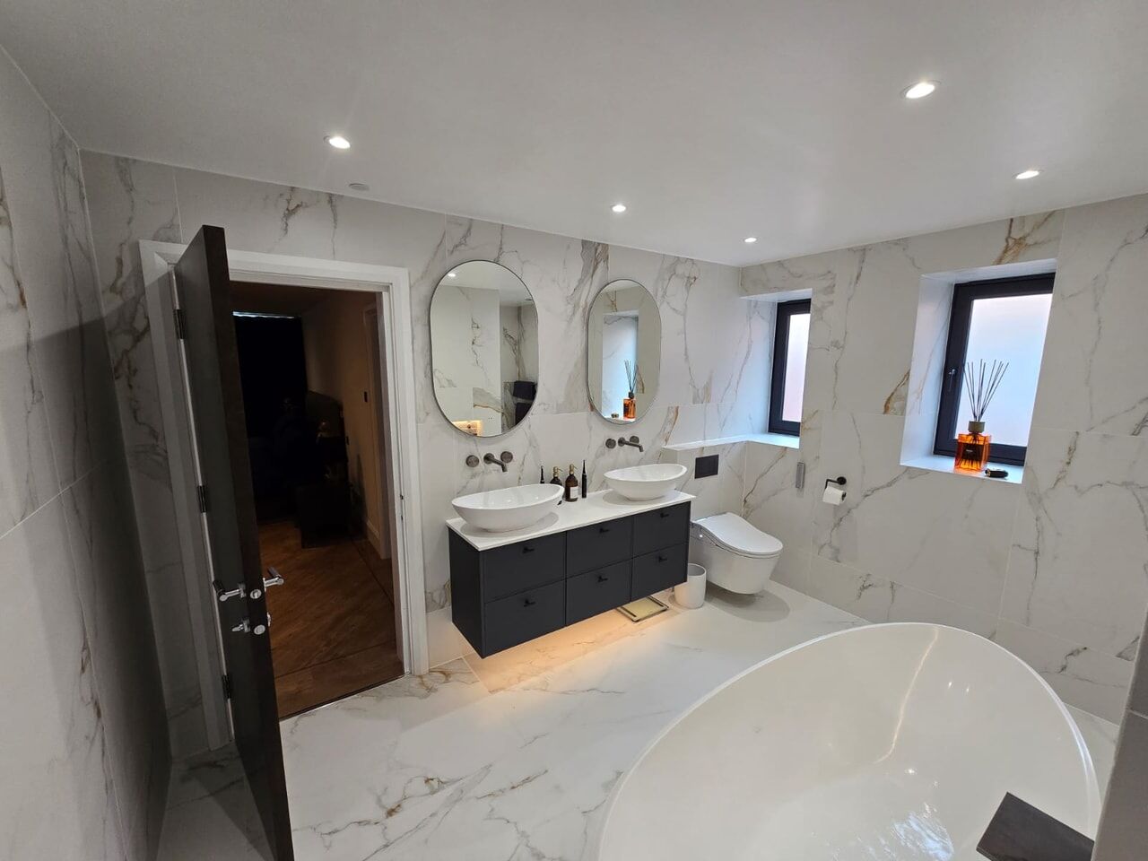 Double vanity with warm underlighting beneath, twin vessel basins and two oval mirrors, a wall-hung WC beyond and a freestanding bath in the foreground, Billericay CM11 master ensuite