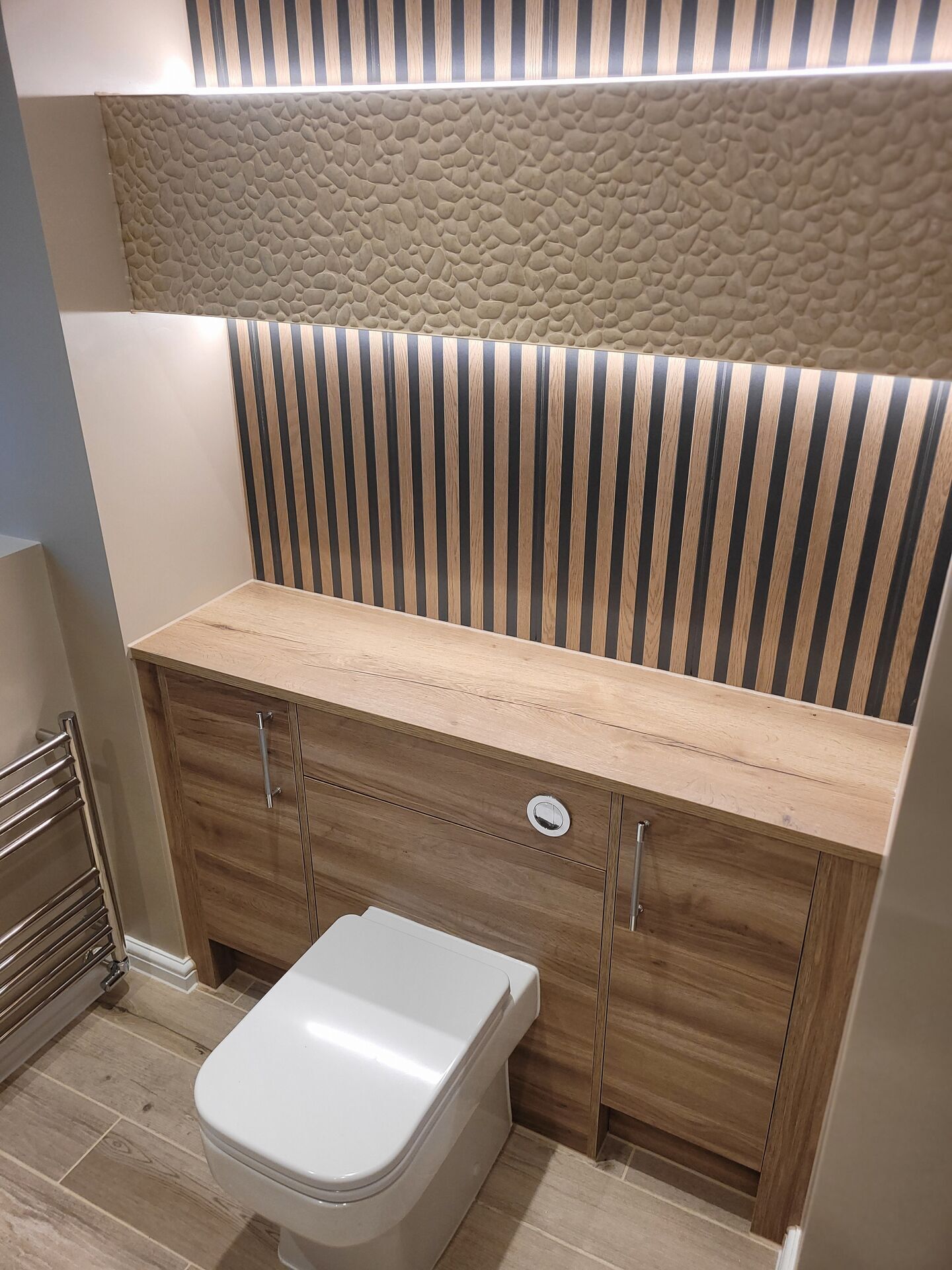 WC tucked into oak-effect fitted furniture below a 3D textured stone-effect strip and a backlit vertical slat wall, Benfleet master ensuite