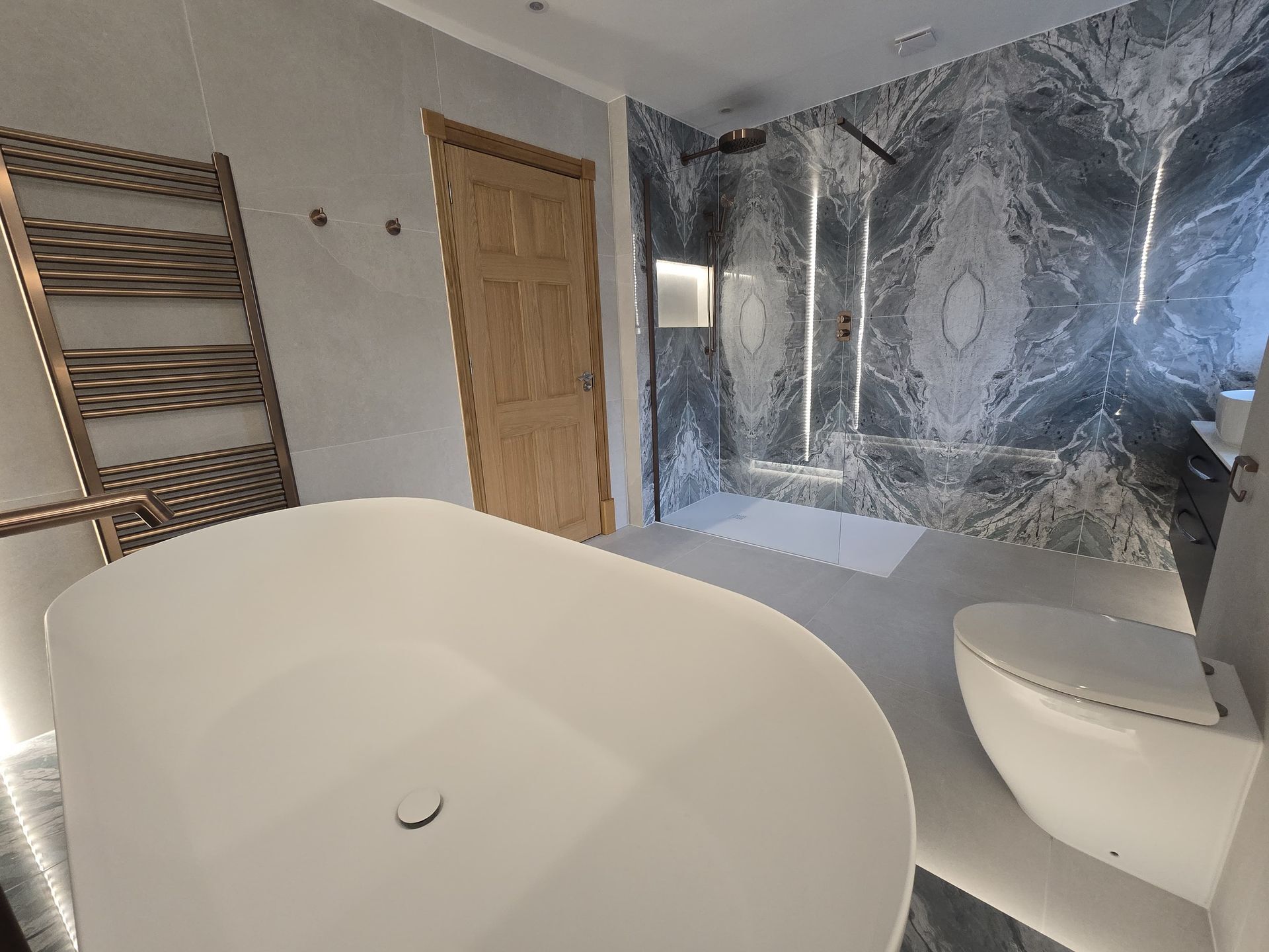 White freestanding bath beside a glass shower and patterned grey wall tiles in a Bradwell bathroom