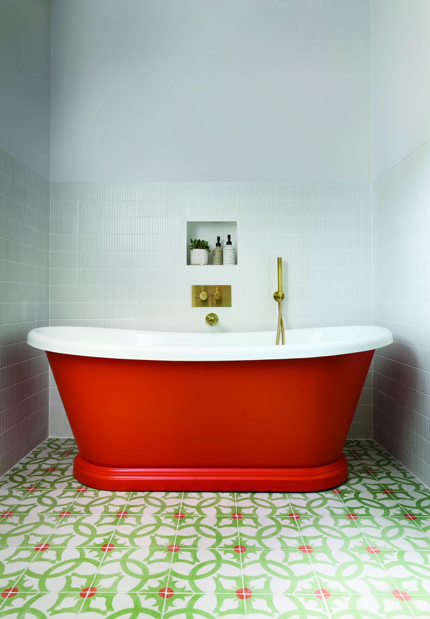 Orange-red painted freestanding bath with pale green walls, brass fittings and patterned green floor tiles