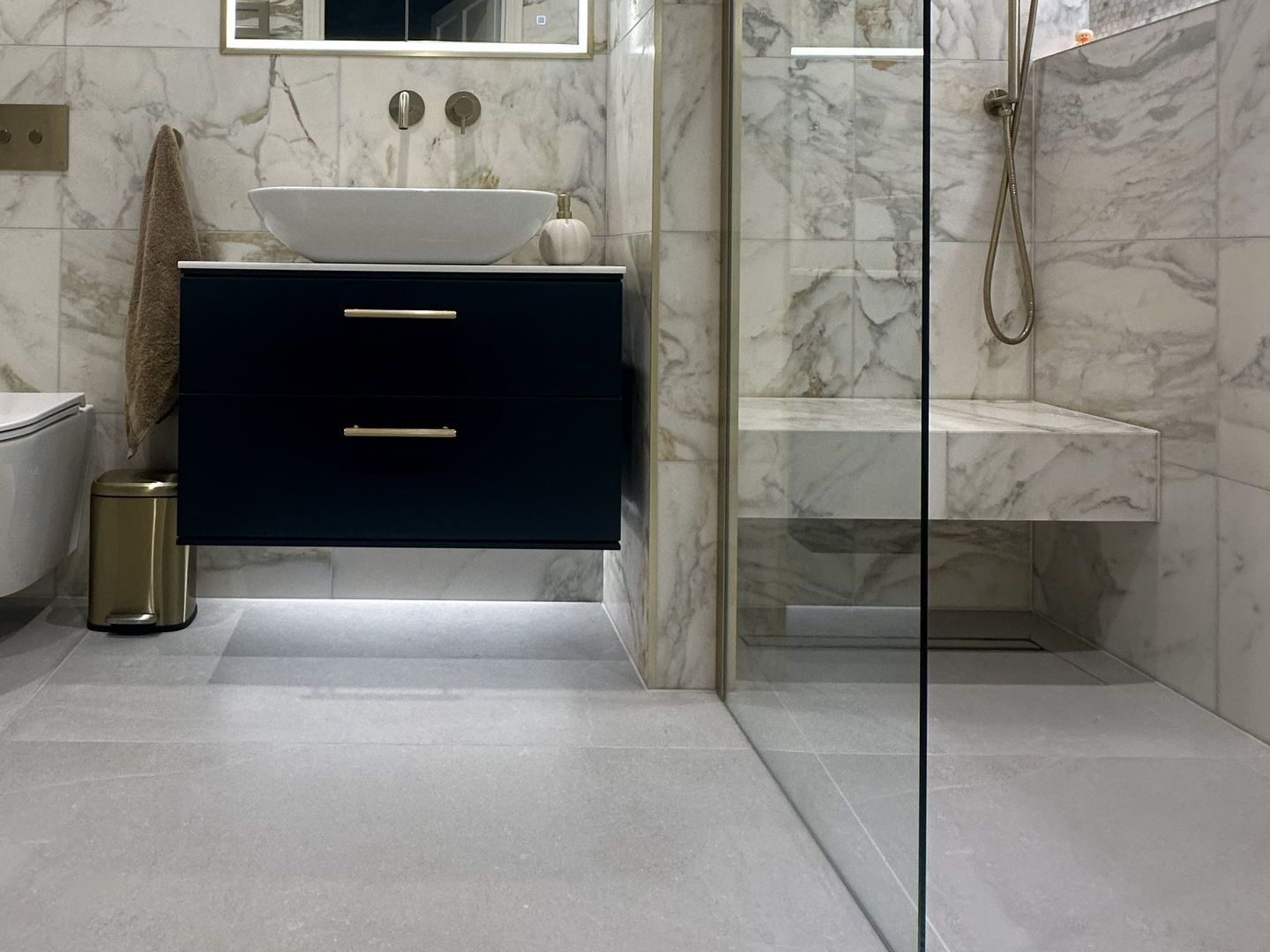 Navy wall-hung vanity beside a glass shower panel, with a thin brass trim on the wall corner and a floating tiled bench inside the shower with no trim on its edge