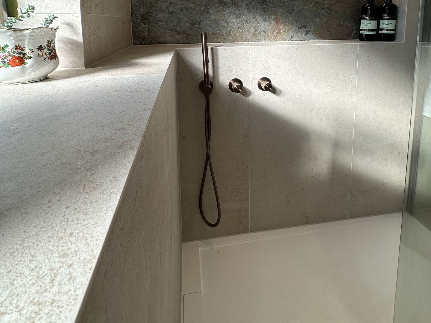 Stone-effect tiled half-wall ledge beside a shower, its top and side meeting on a clean corner with no trim, with a bronze hand shower