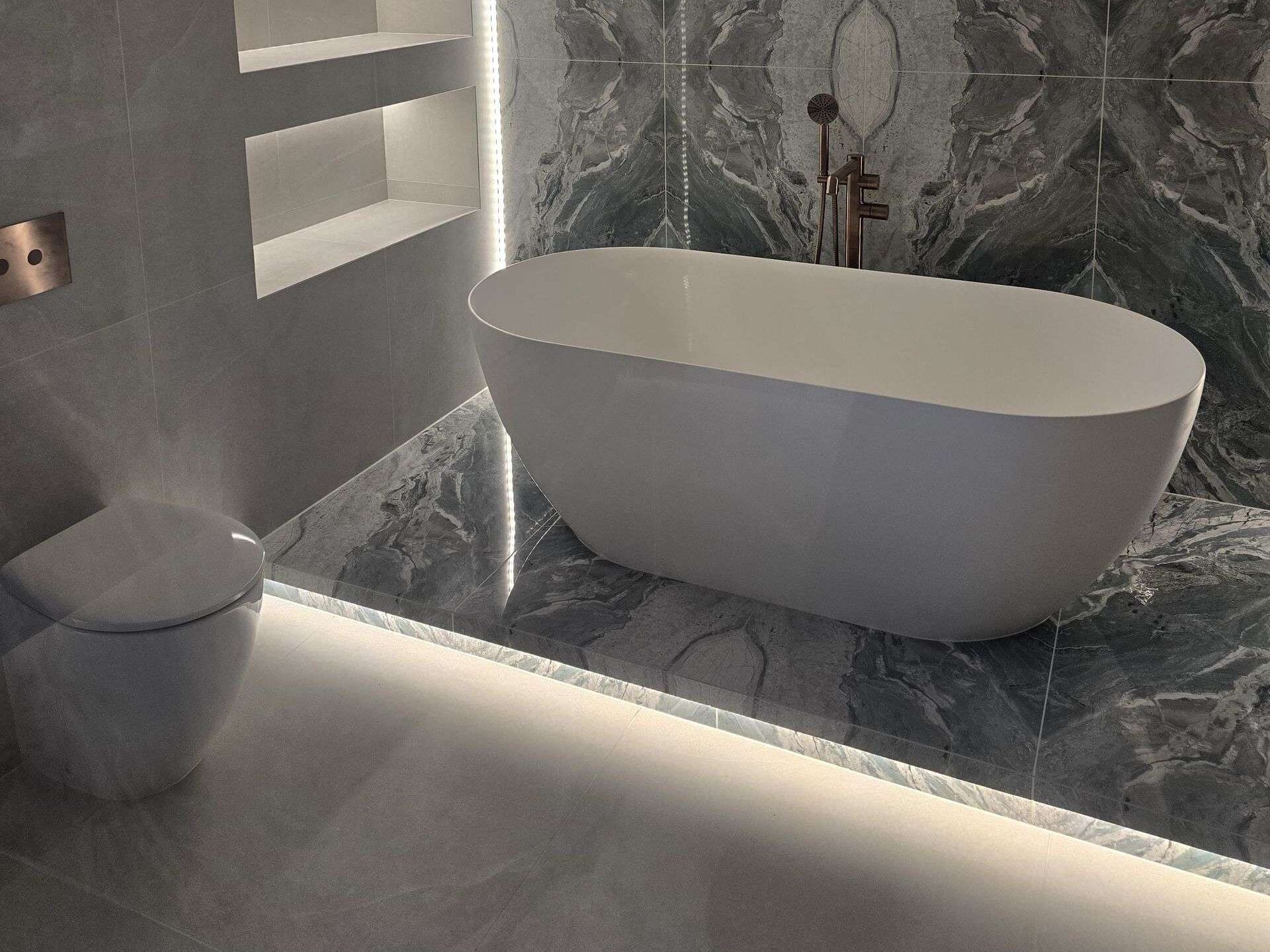Freestanding stone bath on a raised platform tiled in green marble-effect porcelain, its front edge finished with no metal trim and lit from below, in front of a bookmatched feature wall, with two lit niches in the wall beside it.