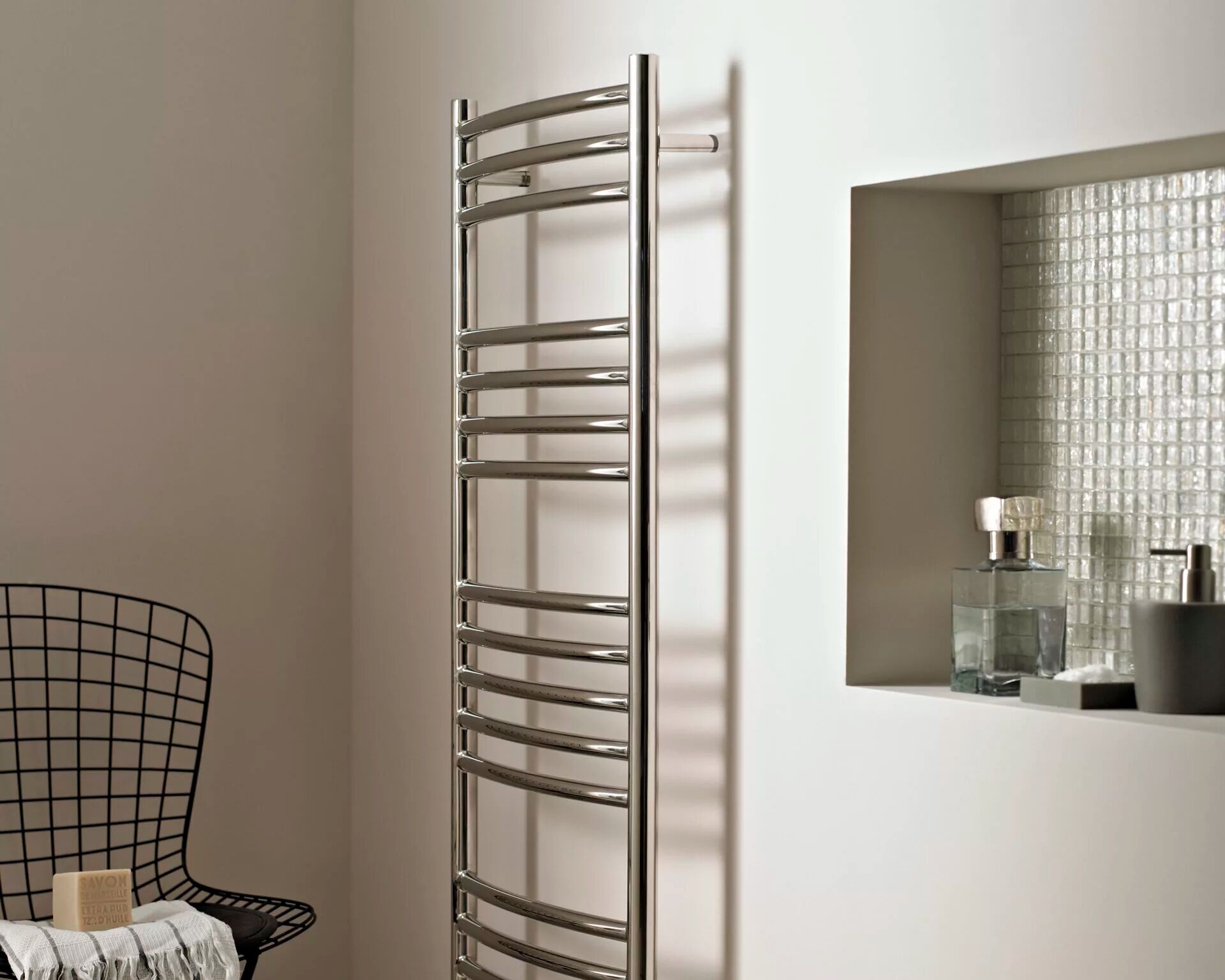 Vogue UK Kerve curved ladder towel rail in polished stainless steel, fixed to a pale wall beside a tiled niche