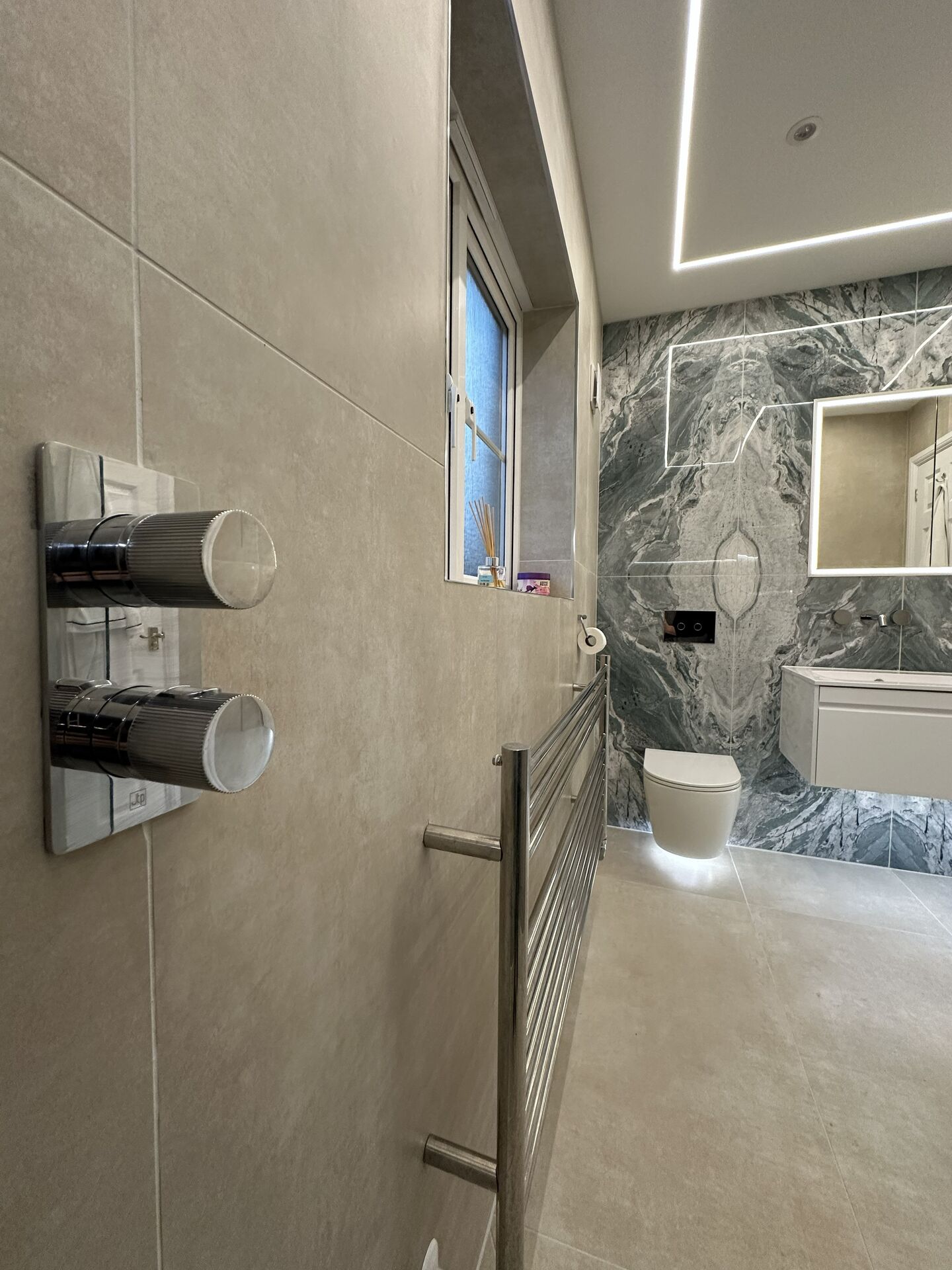 Polished JIS Europe Newick stainless steel towel radiator under the window of a Braintree CM7 wetroom, seen past the shower valve on the same wall
