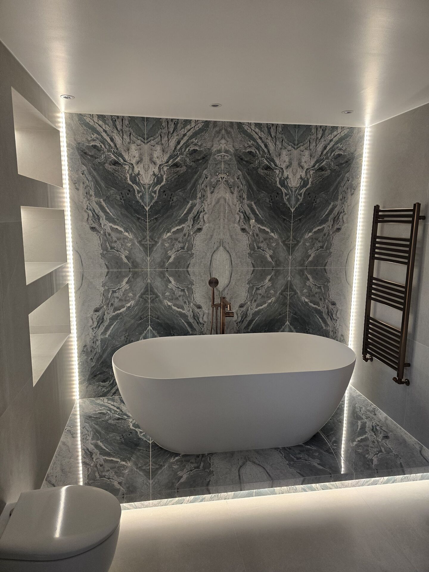 White Crosswater MPRO freestanding stone bath on a raised tiled platform with LED lighting under the platform edge, in front of a bookmatched blue-grey marble-effect feature wall, with a brushed bronze floor-standing bath filler behind the bath