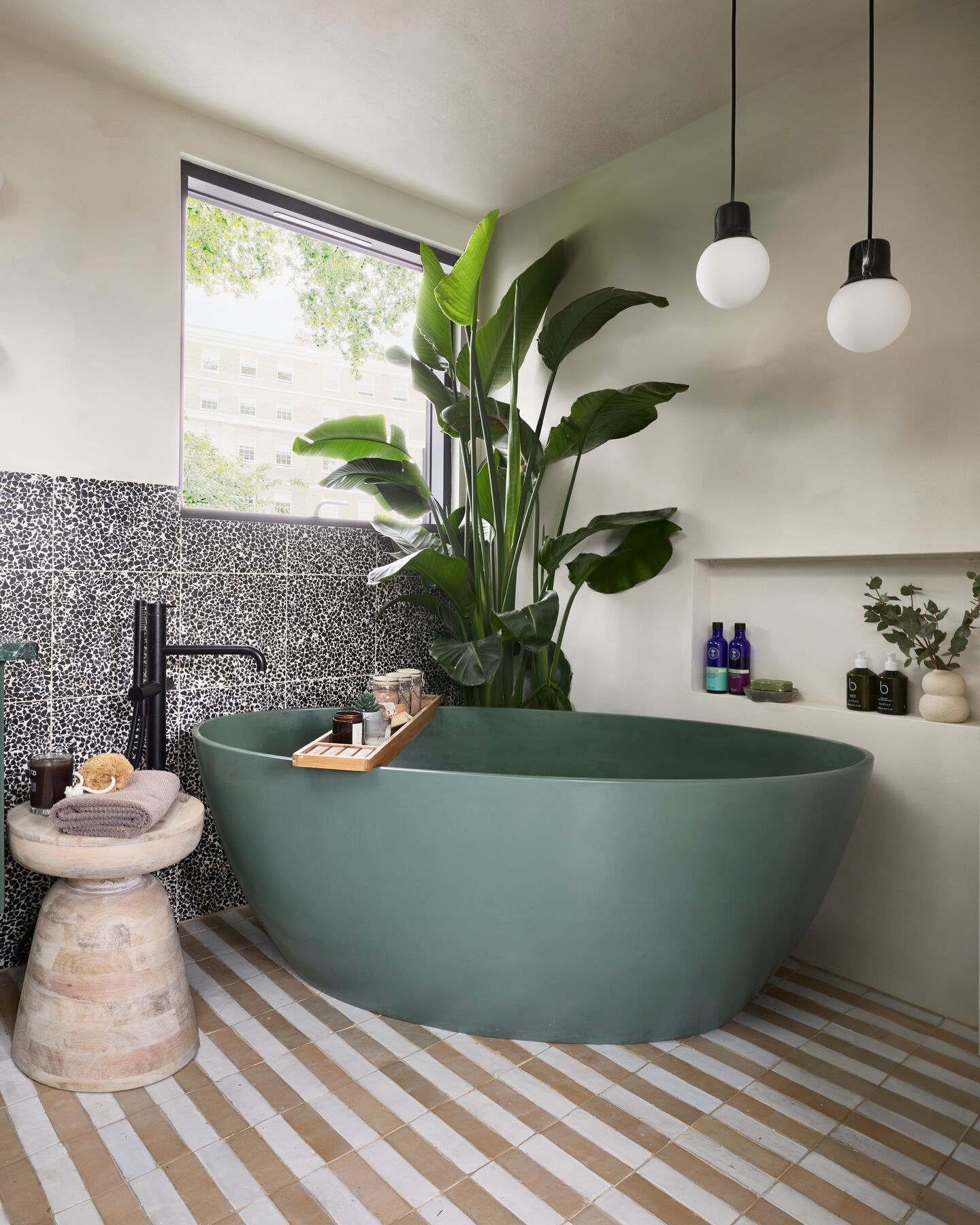 BC Designs Esseta freestanding bath in matt Khaki Green with a black floor-standing bath filler, on a striped tiled floor beside a window, a tall plant and a recessed shelf