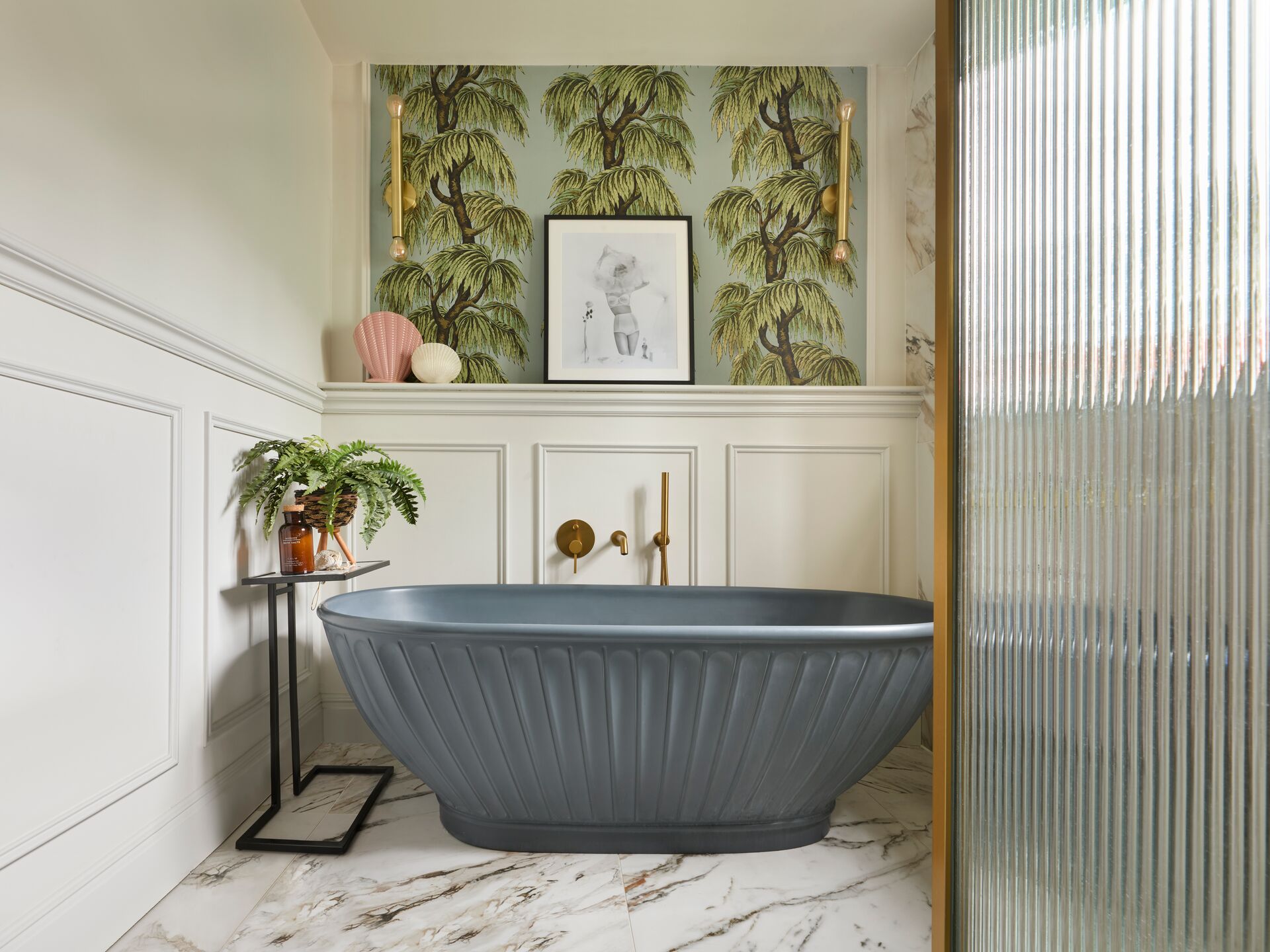 BC Designs Casini freestanding bath in Cian solid surface, finished in the ColourKast colour Powder Blue, with a fluted exterior, standing on a veined marble-look floor in front of white wall panelling and a brass-finish wall-mounted bath filler