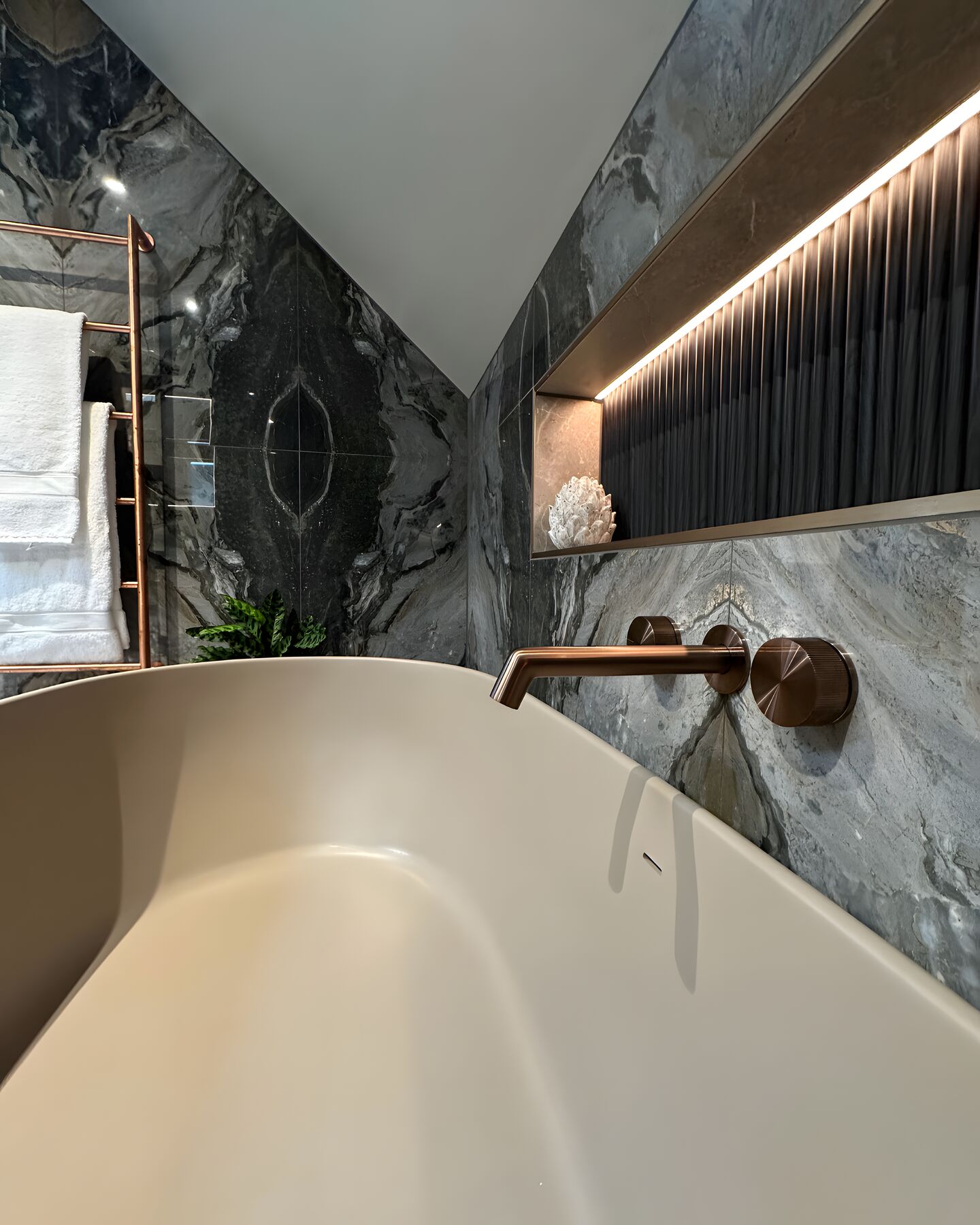 Close view of the matt Light Fawn rim of a BC Designs Crea bath with its overflow slot, a wall-mounted brushed bronze bath filler, dark bookmatched marble-effect wall tiles and an LED-lit ribbed niche above
