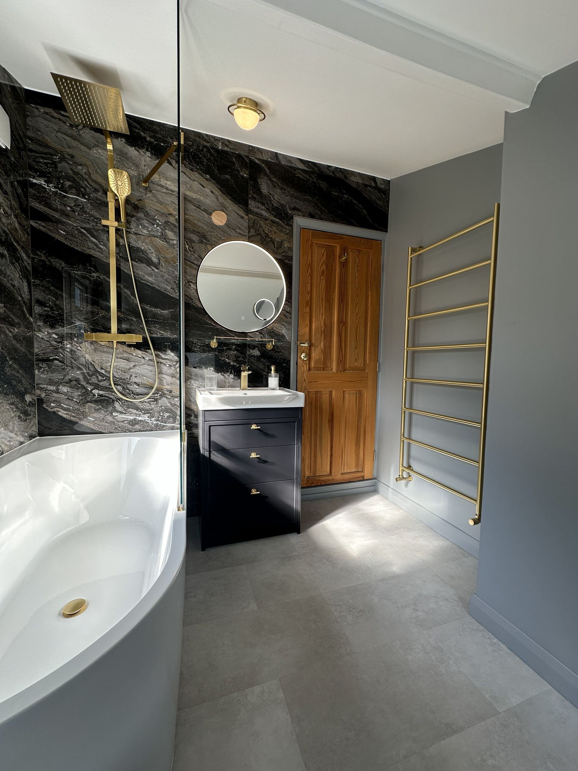 J-shape bath with brushed brass rainfall shower, handset, and glass screen against Ca' Pietra marble feature wall in Braintree CM7