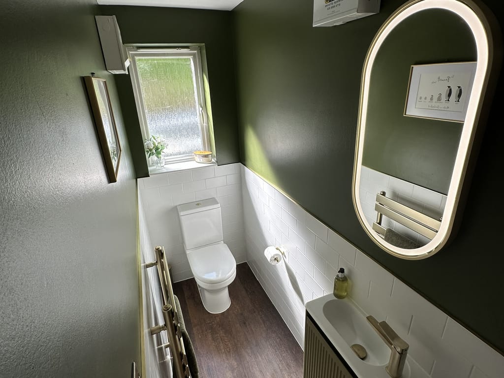 Full cloakroom view with HiB Adorn Olive vanity, Capsule LED mirror, Hix brass radiator, dark green walls and white metro tiles in Great Leighs CM3