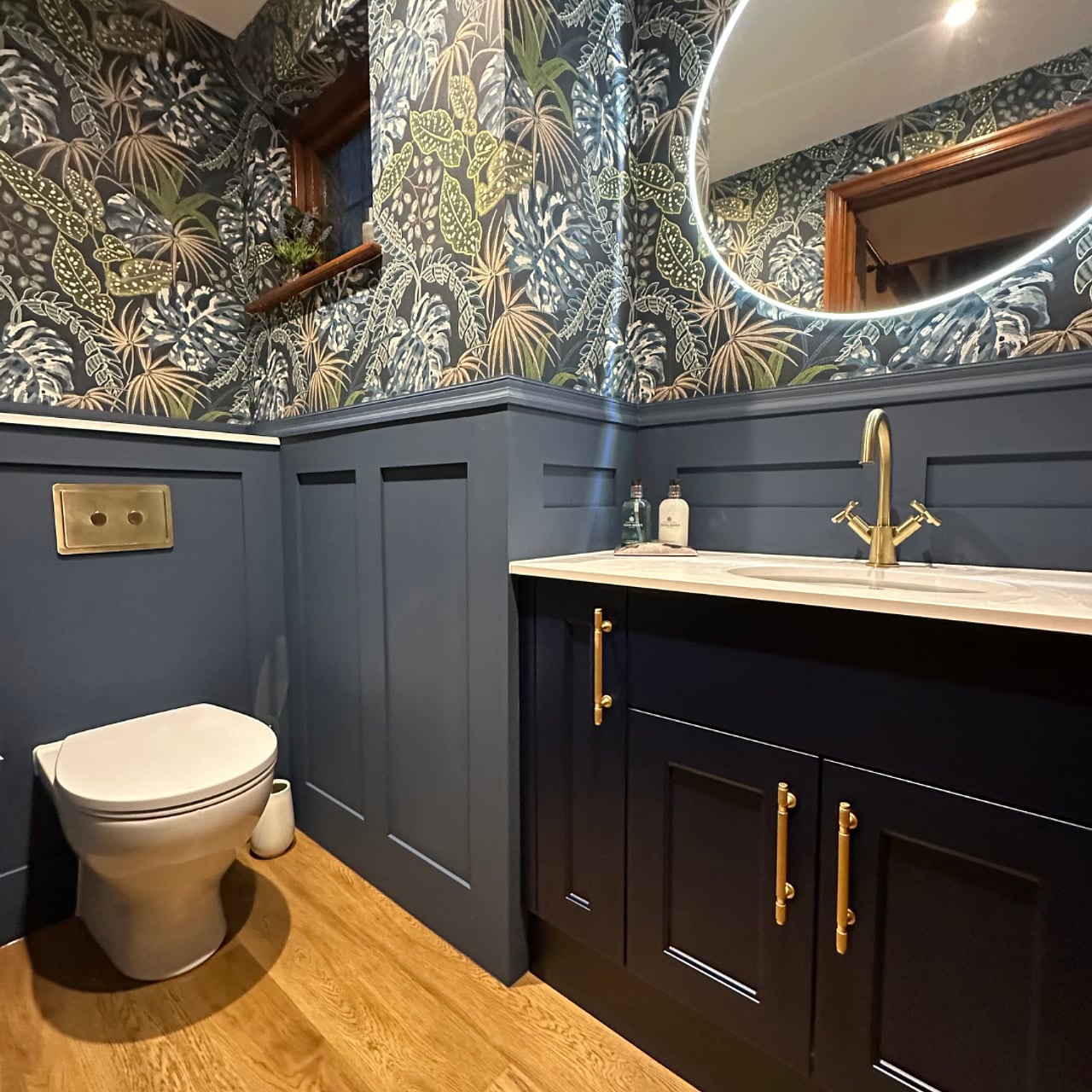 Luxury cloakroom overview with navy Calypso vanity, HiB Air Round mirror, jungle wallpaper above navy panelling, and oak flooring Hatfield Peverel CM3